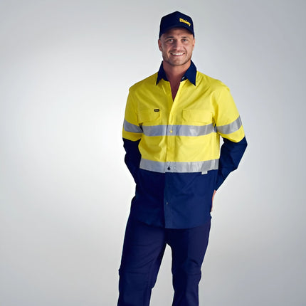 A person stands wearing a long-sleeve work shirt. The shirt is two-toned in bright yellow and dark blue with reflective stripes. They are also wearing a dark blue cap and trousers.