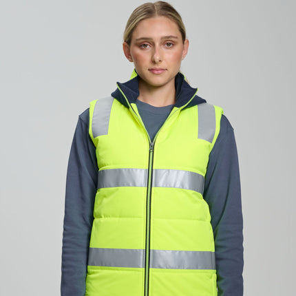 A person stands facing the camera. She wears a neon yellow padded vest with gray reflective stripes over a dark blue long-sleeve shirt. The vest has a dark blue fleece collar and hood.