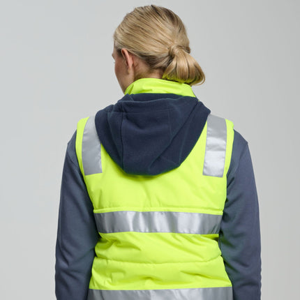 A person stands facing away from the camera. She wears a neon yellow padded vest with gray reflective stripes over a dark gray long-sleeve shirt. The vest has a dark blue fleece hood.