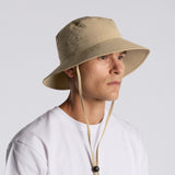 Someone wearing an AS Colour Personalised Nylon Wide‑Brim Bucket Hat in beige, featuring chin straps for sun protection, and a white t-shirt, faces slightly right against a plain light background.
