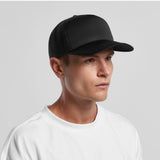 A person in a white t-shirt and an AS Colour Frame Foam Trucker Cap looks to the side against a neutral gray background.