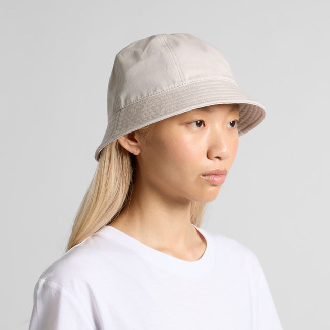 A person with long, straight blonde hair wears an AS Colour Personalised Wo’s Brim Bucket Hat in light beige and a plain white t-shirt, facing slightly right against a neutral gray background.