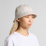 A person with long, straight blonde hair wears an AS Colour Personalised Wo’s Brim Bucket Hat in light beige and a plain white t-shirt, facing slightly right against a neutral gray background.