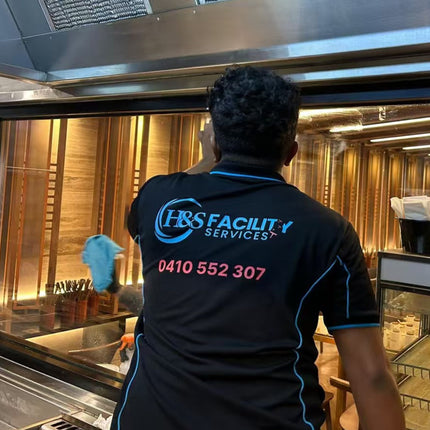Wearing a My Promo Shop JB’s Wear Arachnid Polo with “0410 552 307,” a worker cleans glass in an upscale, warmly lit restaurant. The breathable polo delivers comfort and professional flair while on the job.