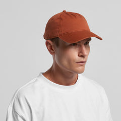 A person wears a plain white T-shirt and an AS Colour Access Five Panel Cap in rust, looking slightly to the right against a neutral gray background.