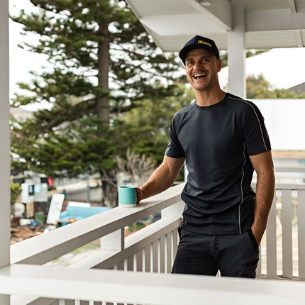 A man is smiling and holding a mug while standing on a porch. He wears a dark gray short-sleeve shirt with white piping, dark shorts, and a dark blue cap.