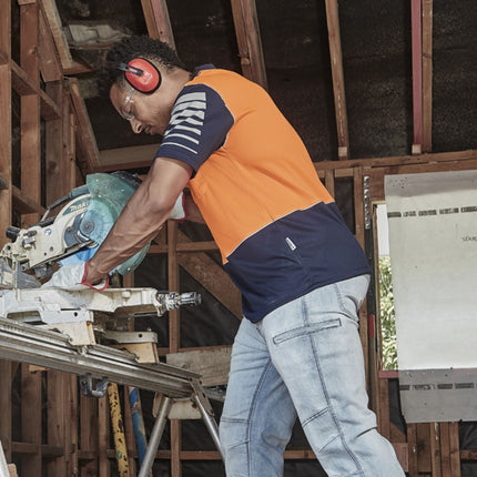 A man cuts wood with a saw. He wears an orange and dark blue polo with reflective stripes, jeans, and ear protection.