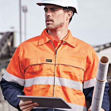 A man in a bright orange and dark blue work jacket holds a tablet and blueprints. The jacket has reflective stripes and he wears a white hard hat.
