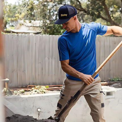 A person stands outdoors, shoveling. He wears a blue short-sleeve shirt, beige pants, and a dark blue cap with a yellow logo.