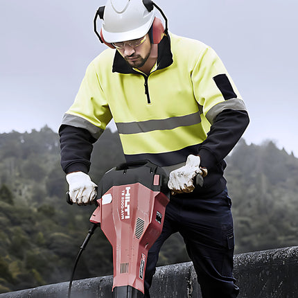 A construction worker uses a jackhammer. He is wearing a neon yellow and dark blue high-visibility work sweater, a white hard hat, and safety glasses and gloves.