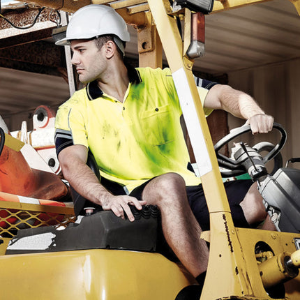 A worker wearing a neon yellow high-visibility polo shirt with dark blue accents drives a forklift. He is wearing a white hard hat.