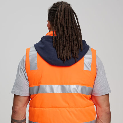 A person stands facing away from the camera. They are wearing an orange padded vest with gray reflective stripes over a gray t-shirt. The vest has a dark blue fleece hood.