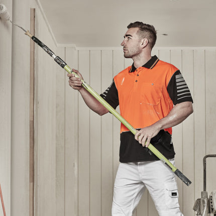  A man paints a wall. He wears an orange and black polo with reflective stripes and holds a roller on a long pole.