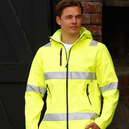 A man outdoors wears a neon yellow work jacket with gray reflective stripes. He has one hand in a pocket and is looking to the side.