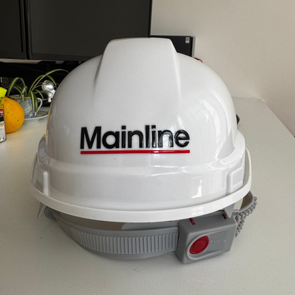 A white hard hat with the Mainline logo printed on the front, resting on a light-colored desk with a computer monitor in the background.