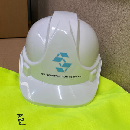 A glossy white construction hard hat with a blue triangular logo and the text 'A2J CONSTRUCTION SERVICES' resting on a bright yellow high-visibility safety vest. The background shows a cardboard box and a textured brown surface.