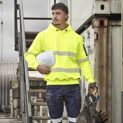 A person walks down stairs. He is wearing a neon yellow hoodie with reflective stripes and a gray zippered sleeve pocket. He is holding a white hard hat and work gear.