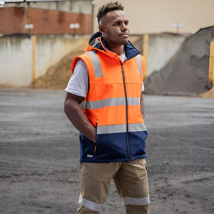 A person stands outdoors wearing a two-tone work vest. The vest is orange and dark blue with gray reflective stripes. He has his hands in his pockets.