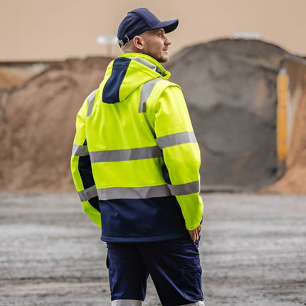 A person stands with his hands in his pockets, wearing a neon yellow and dark blue work jacket. The jacket has a hood and gray reflective stripes.
