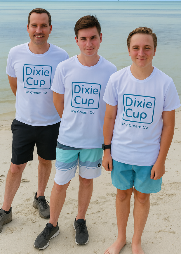 Three people wearing matching white "Dixie Cup Ice Cream Co." t-shirts stand on a sandy beach with calm ocean water in the background. The individuals are smiling and dressed in casual beachwear, with two wearing shorts and one in sneakers.