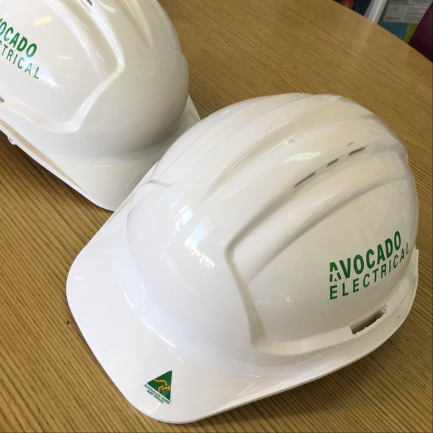 Two white hard hats with 'Avocado Electric' branding on a wooden surface.