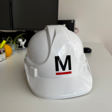 A glossy white hard hat with a black letter 'M' and a red underline printed on the front, displayed on a white desk in an office setting.