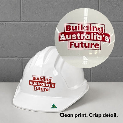 A white hard hat with a red and white 'Building Australia's Future' slogan printed on it, shown against a gray brick wall background with a circular inset showing a close-up of the print. Text at the bottom reads 'Clean print.