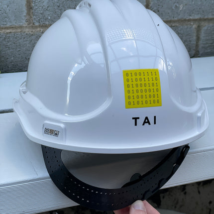 A white hard hat with a black chin strap resting on a white metal rail, with a hand holding the strap. The hard hat has a yellow sticker with binary code and the brand name 'TAI' printed on it.
