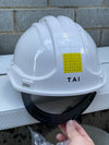 A white hard hat with a black chin strap resting on a white metal rail, with a hand holding the strap. The hard hat has a yellow sticker with binary code and the brand name 'TAI' printed on it.