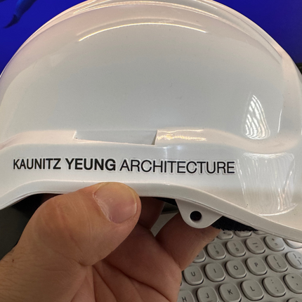 A white hard hat with 'KAUNITZ YEUNG ARCHITECTURE' printed in black on the side, held by a hand in front of a computer keyboard and monitor.