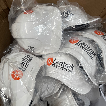 A pile of white hard hats wrapped in clear plastic inside a cardboard box. Several hats show the printed logo 'Lentrak Electrical' in black with an orange circular emblem.