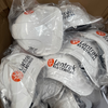 A pile of white hard hats wrapped in clear plastic inside a cardboard box. Several hats show the printed logo 'Lentrak Electrical' in black with an orange circular emblem.