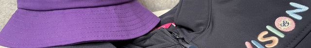 A purple bucket hat and a dark gray zip-up hoodie, both featuring the word 'VISION' embroidered in pastel multicolor letters with a small smiley face inside the letter 'O'.