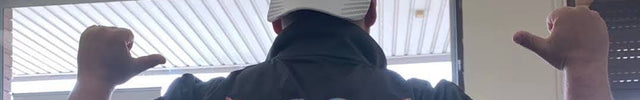 A man stands with his back to the camera indoors, wearing a black long-sleeve work shirt with a large white printed back design, teal sleeves, white shorts, and a white cap. The shirt has visible text and logos including Dulux and 2nd Century Painting and House Improvements.