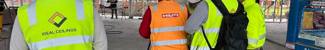 A group of construction workers wearing high-visibility safety vests and white hard hats gathered under a ceiling structure on a construction site. One worker on the left wears a bright yellow-green vest with the brand name 'IDEAL CEILINGS' printed on the back.