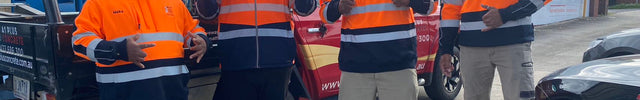 Four workers standing outdoors next to a red work truck, wearing matching orange and navy blue hi-vis hoodies with reflective stripes, caps, and work boots.