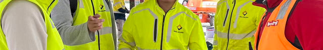 A group of five construction workers standing on a job site wearing hard hats. Four workers are wearing bright yellow hi-vis jackets, and one is wearing an orange hi-vis vest.