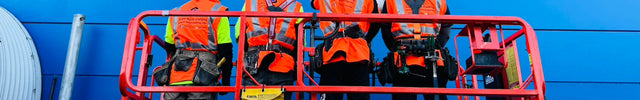 Four workers wearing bright orange hi-vis shirts with reflective tape and hard hats, standing in a red aerial work platform against a blue building