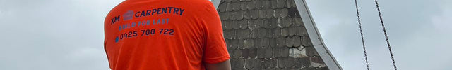 A construction worker viewed from behind wearing a bright orange hi-vis t-shirt with a custom carpentry logo, a tool belt, and a white hard hat, looking at a building under construction.