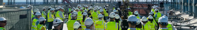 A large group of construction workers wearing high-visibility yellow safety vests and white hard hats gather on a rooftop under a clear blue sky, with a red carpet leading up the slope toward a steel structure.