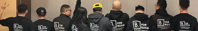 A group of ten people standing with their backs to the camera in a conference room, wearing matching black t-shirts and hoodies with 'B FOR BUILD' printed in white on the back.