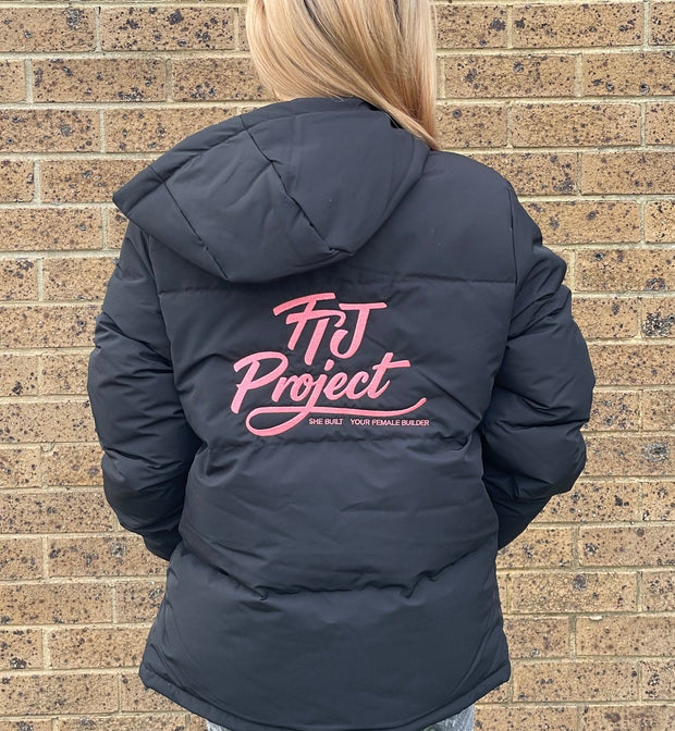 A person viewed from behind wearing a dark navy puffer jacket with a pink 'FIT Project' logo printed on the back, standing against a tan brick wall.