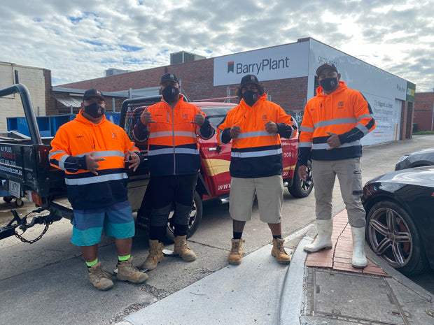 Four men wearing high-visibility orange and navy work jackets, face masks, and boots stand outdoors in front of a red truck and a building with a large 'BarryPlant' sign.