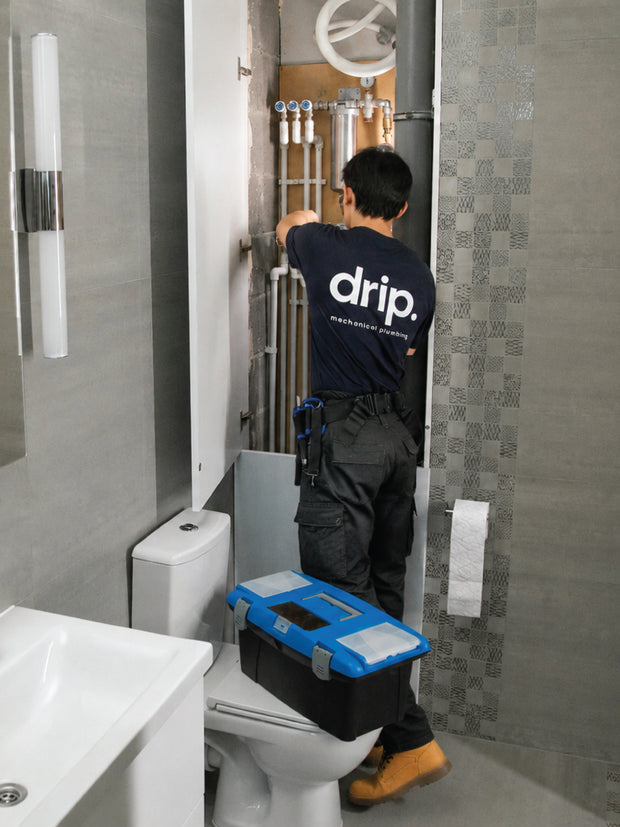 A plumber wearing a navy blue 'drip.' work shirt and dark cargo pants stands in a modern bathroom, working on plumbing fixtures behind a toilet.