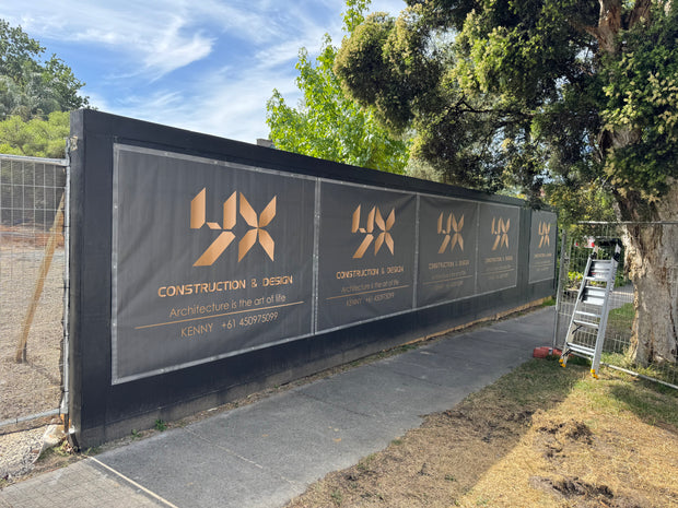 A long black construction hoarding panel with gold logo and text for 'UX Construction & Design' installed outdoors along a sidewalk beside trees and a ladder.