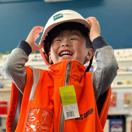 A young boy smiling and laughing while wearing a white construction hard hat and an orange safety vest with reflective stripes in a retail store setting.